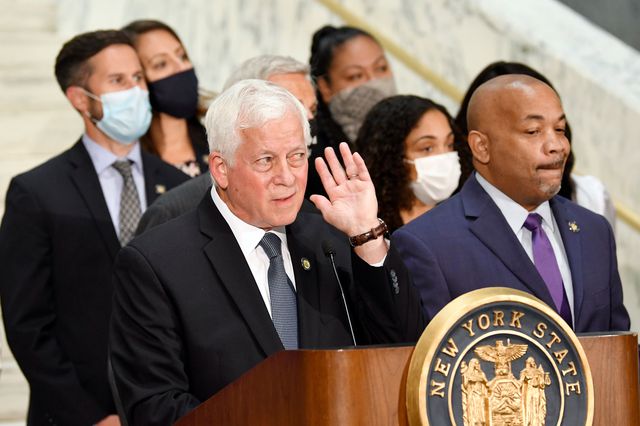 Assembly Judiciary Committee chair Charles Lavine, who has white hair, listens, with his hand his ear, during a press conference. He is unmasked, as is ASsembly Speaker Carl Heastie, who is leading next to him at a lectern. Other masked members of the Judiciary Committee stand behind them
